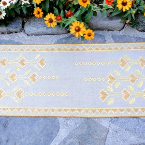 Può includere: Un tessuto rettangolare grigio chiaro con ricami gialli intricati. Il design presenta motivi floreali e geometrici, con un bordo dettagliato. Il tessuto è probabilmente un tappeto o un runner decorativo.