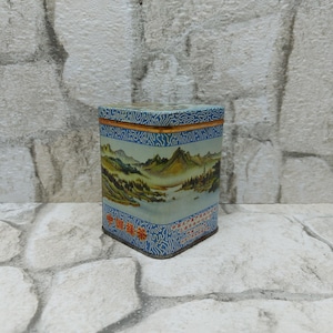 May include: Square tin box with a blue and white design featuring a landscape scene with mountains, trees, and water. The box has a gold trim and Chinese characters on the front.