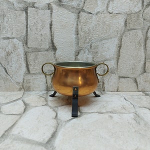 Puede incluir: Un caldero de color cobre con dos asas de latón y tres patas negras. El caldero tiene un interior plateado y está colocado sobre un fondo de pared de piedra. Esta pieza decorativa es adecuada para la decoración del hogar.