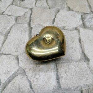 May include: A heart-shaped brass box with a shiny, golden finish. The box has a small, circular detail on the top and a slight indentation on the front. The object is placed on a light gray stone surface.