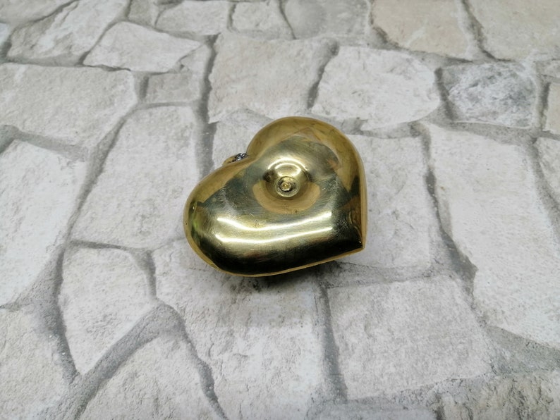 May include: A heart-shaped brass box with a shiny, golden finish. The box has a small, circular detail on the top and a slight indentation on the front. The object is placed on a light gray stone surface.