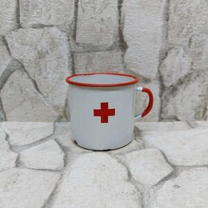 May include: A white enamel mug featuring a red cross, red rim, and handle. The mug is positioned against a textured stone backdrop, evoking a vintage or medical aesthetic. The design of the mug is straightforward and practical.
