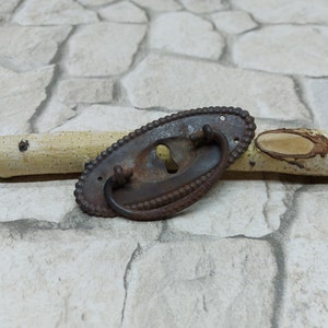 May include: A rusty metal oval-shaped drawer pull with a beaded edge and a keyhole. The pull has a curved handle and is on a light gray stone background.