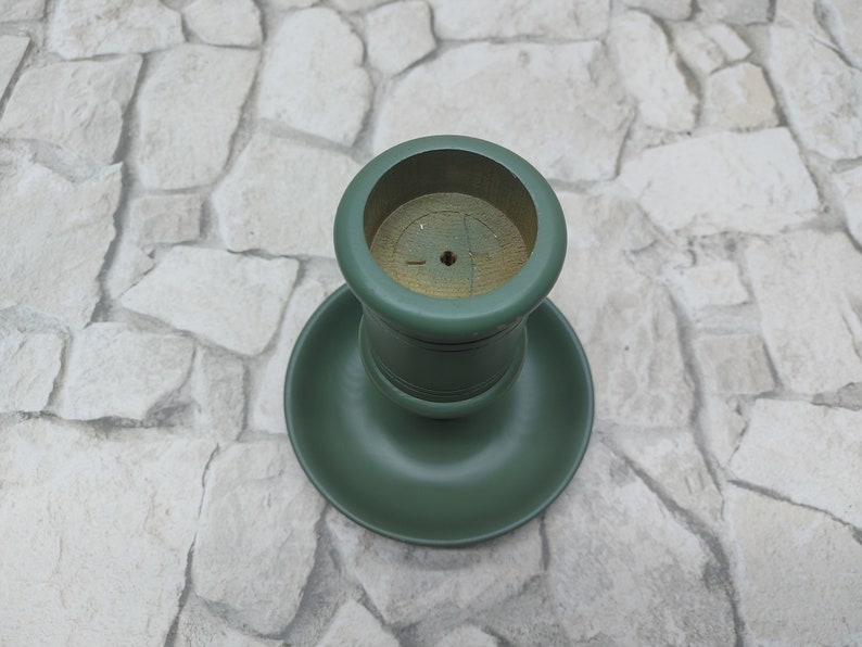 May include: A green wooden candlestick holder. It has a circular base and a cylindrical top. The inside of the top is natural wood. The holder is painted in a matte green finish.