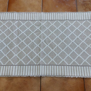 Puede incluir: Camino de mesa beige y blanco con estampado de diamantes. El camino de mesa tiene un patrón de diamantes repetido en blanco sobre un fondo beige. Los bordes presentan un diseño a rayas en beige y blanco, con un borde festoneado. Este camino de mesa es adecuado para la decoración del hogar.