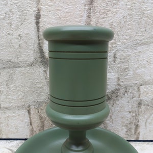 May include: A sage green wooden candlestick holder. The holder has a round base and a cylindrical top, with decorative horizontal lines. The finish is smooth and matte. The background is a textured, light-coloured wall.