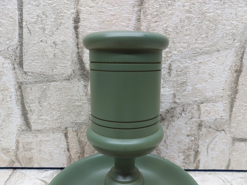 May include: A sage green wooden candlestick holder. The holder has a round base and a cylindrical top, with decorative horizontal lines. The finish is smooth and matte. The background is a textured, light-coloured wall.