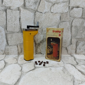 May include: A yellow manual coffee grinder with a black handle and a silver top, next to its original box. The box is labeled "tramp" and features an illustration of the grinder. Several coffee beans are scattered in front.
