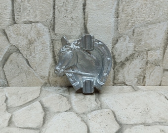 May include: Silver-colored ashtray with a horse head design. The ashtray features a horseshoe shape with a horse head in the center and two built-in rests for cigarettes. The ashtray is made of metal and has a rustic appearance.