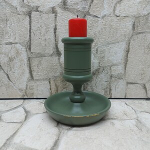 May include: A green wooden candlestick with a red candle. The candlestick has a round base and a central stem with a circular top to hold the candle. The candle is a short, cylindrical shape. The candlestick is painted green with black accents.