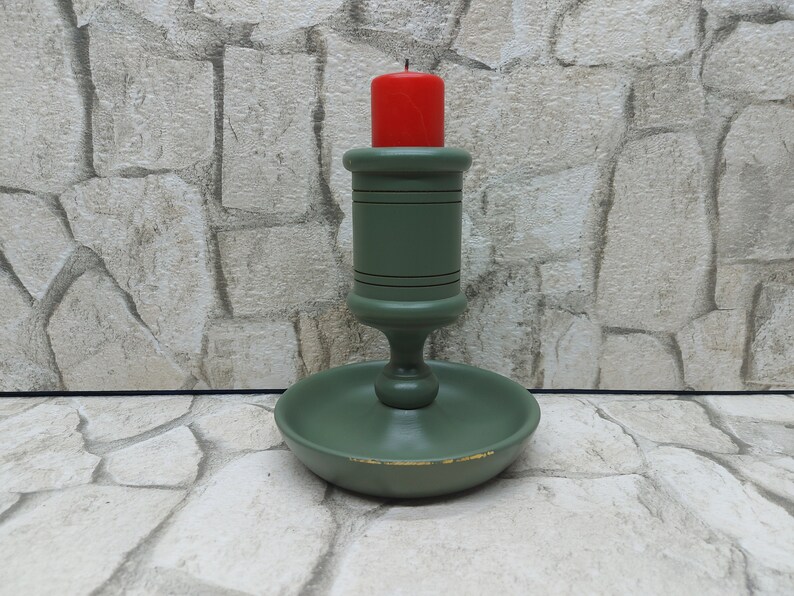 May include: A green wooden candlestick with a red candle. The candlestick has a round base and a central stem with a circular top to hold the candle. The candle is a short, cylindrical shape. The candlestick is painted green with black accents.
