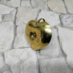 May include: A heart-shaped brass container with a small handle. The container has a smooth, polished surface and a small, circular indentation on the front. The brass has a warm, golden color. The container is likely used for decorative purposes.