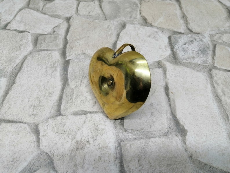 May include: A heart-shaped brass container with a small handle. The container has a smooth, polished surface and a small, circular indentation on the front. The brass has a warm, golden color. The container is likely used for decorative purposes.