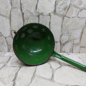 May include: A vintage green enamel colander with a long handle. The colander has a round bowl with numerous small holes for draining. The handle is attached to the side of the bowl. The colander is a kitchen utensil.