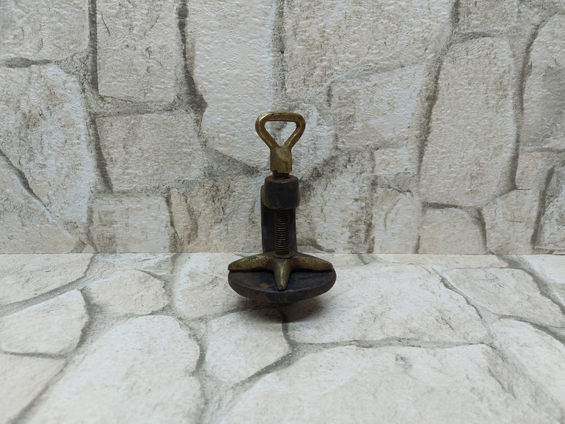 May include: A vintage metal clamp with a brass handle and screw mechanism. The clamp has a dark, aged appearance, with a star-shaped base and a threaded rod. The handle is a loop shape.