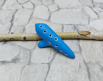 May include: A bright blue ceramic ocarina with multiple finger holes. The wind instrument is oval-shaped with a mouthpiece and sits on a light-colored wooden stick against a stone background. A musical instrument.