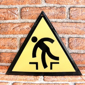 Puede incluir: Una señal de advertencia triangular amarilla con borde negro. La señal tiene una silueta negra de una persona que tropieza con un escalón. La señal dice "Precaución - Escalón"