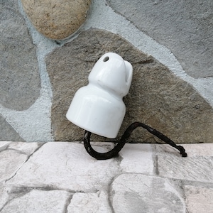 May include: A white ceramic insulator with a curved, dark metal wire attached. The insulator has a rounded top with a hole and a curved shape. The background is a stone wall and a stone floor.