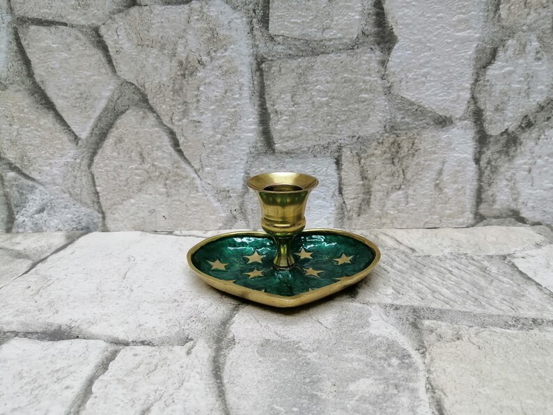 May include: A brass candlestick holder with a heart-shaped base. The base is green with gold stars and a gold border. The candlestick holder is made of brass.