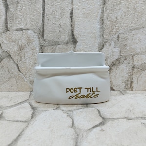 Può includere: Un portacorrispondenza in ceramica bianca con apertura rettangolare e parte superiore curva. Le parole "POST TILL table" sono stampate in lettere dorate sul davanti. Il portacorrispondenza ha un aspetto leggermente sgualcito.