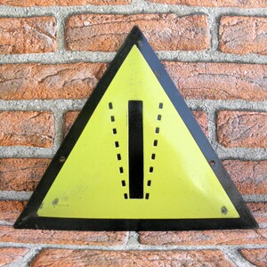 May include: A yellow triangular warning sign with a black border and a black vertical line with dashed lines on either side. The sign is made of metal and has a distressed look.