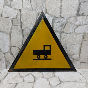 May include: A yellow triangular warning sign with a black border. The sign has a black silhouette of a truck with wheels on a track.