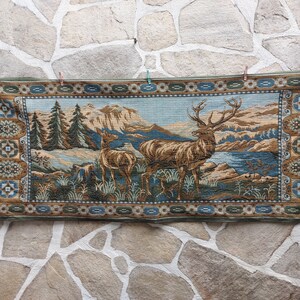 Pode incluir: Uma tapeçaria com uma paisagem com dois veados, um macho e uma fêmea, perto de um lago. A tapeçaria tem um esquema de cores azul, castanho e verde, com uma borda estampada. O fundo inclui árvores e montanhas.
