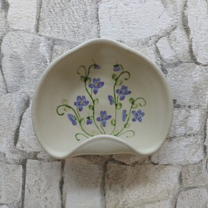 Puede incluir: Apoyacucharas de cerámica blanquecina con diseño floral. El apoyacucharas tiene una forma curvada con un pequeño labio y está decorado con flores moradas y tallos verdes. El fondo es un patrón de piedra gris claro.