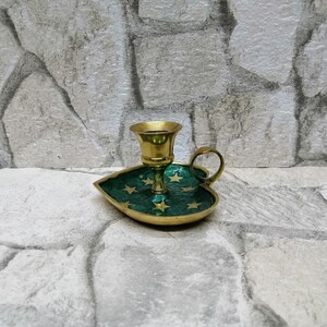 May include: A brass candle holder with a green leaf-shaped base. The base is decorated with gold stars. The candle holder has a small handle and a cup for the candle. The background is a stone wall.