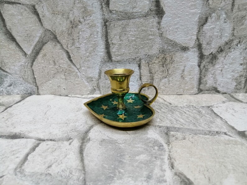 May include: A brass candle holder with a green leaf-shaped base. The base is decorated with gold stars. The candle holder has a small handle and a cup for the candle. The background is a stone wall.