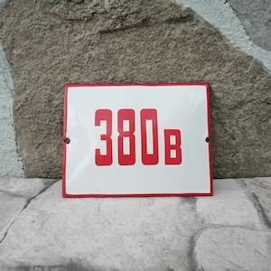 May include: A rectangular white metal sign with a red border, displaying the number "380B" in red block letters. The sign has two mounting holes and is set against a stone background.