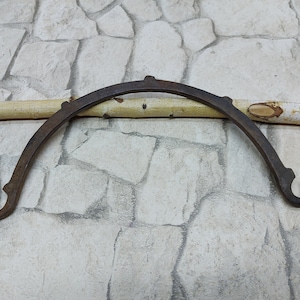 Peut inclure: Un support en métal brun foncé incurvé avec des extrémités décoratives et de petites protubérances sur le dessus. Le support est arqué et semble être forgé à la main. Une tige en bois de couleur claire repose derrière le support, sur un fond de pierre.