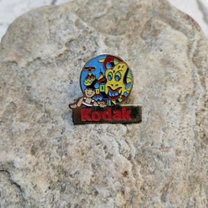 Puede incluir: Un pin de color dorado con una imagen de dibujos animados de colores de una persona y un monstruo. El pin tiene la palabra "Kodak" en letras rojas sobre un fondo dorado.