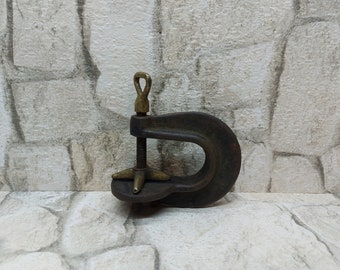 May include: A vintage C-clamp made of dark metal with brass accents. The clamp has a threaded rod for adjusting the opening and a loop for hanging. The background is a textured, light-coloured wall.