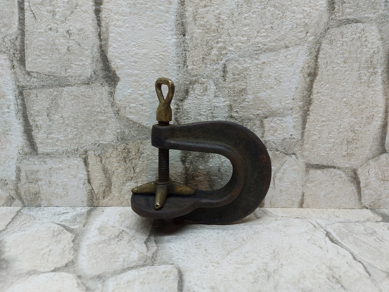 May include: A vintage C-clamp made of dark metal with brass accents. The clamp features a threaded rod for adjusting the opening and a loop for hanging. The background is a textured, light-colored wall.