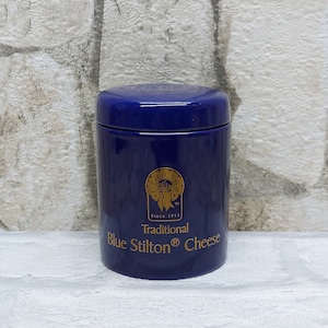 May include: A cobalt blue ceramic jar with a matching lid. The jar features gold lettering that reads "Traditional Blue Stilton Cheese" and a gold emblem. The jar is cylindrical and sits on a white surface.