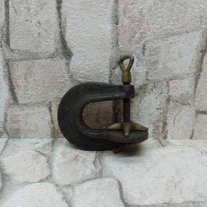 May include: A vintage C-clamp with a dark, weathered appearance. The clamp features a curved, U-shaped frame, a threaded rod, and a brass handle and adjustment components. The tool is designed for securing objects during woodworking or other projects.