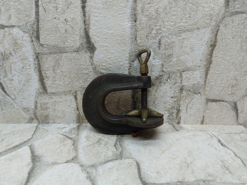 May include: A vintage C-clamp with a dark, weathered appearance. The clamp features a curved, U-shaped frame, a threaded rod, and a brass handle and adjustment components. The tool is designed for securing objects during woodworking or other projects.