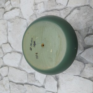 May include: A green, round wooden object with a flat base. The base has the text "L. Karlsson Stiffz 1974" written in black script. The object is painted in a muted green colour.