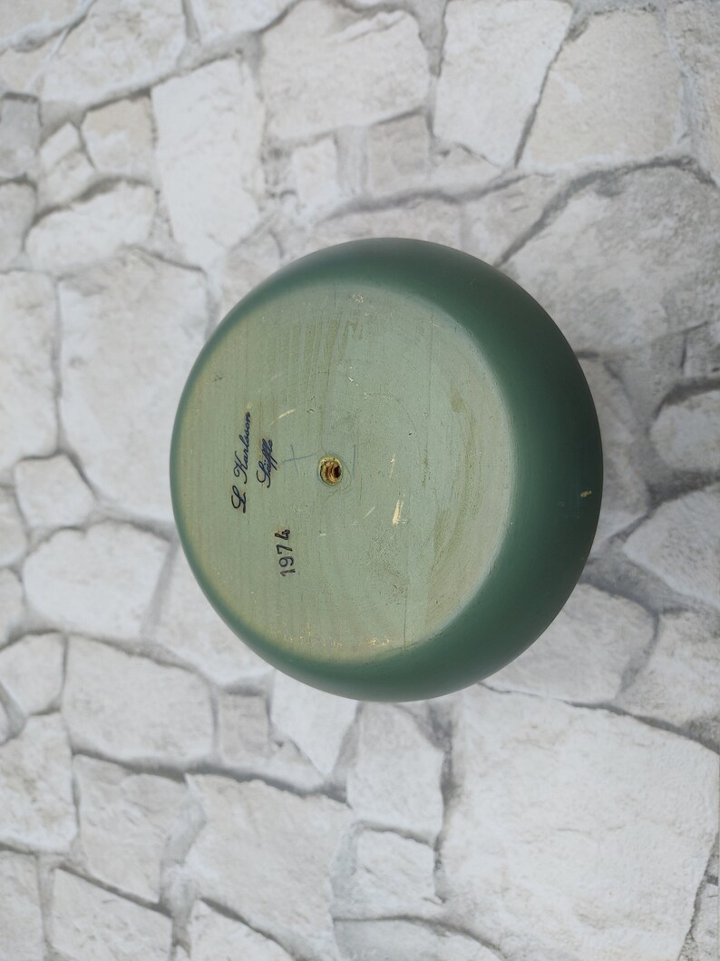 May include: A green, round wooden object with a flat base. The base has the text "L. Karlsson Stiffz 1974" written in black script. The object is painted in a muted green colour.