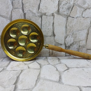 Puede incluir: Una sartén de latón para caracoles con siete hendiduras circulares. Tiene un mango de madera y un pequeño lazo para colgar. La superficie de latón tiene una textura martillada. La sartén está diseñada para cocinar y servir caracoles.