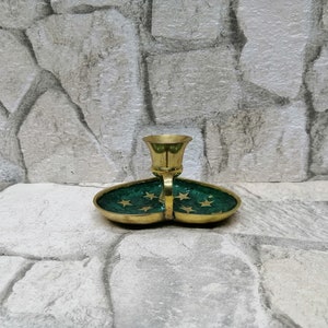 May include: A brass candle holder with a green enamel base. The base is shaped like two connected ovals and is decorated with gold stars. The candle holder is on a stone surface.