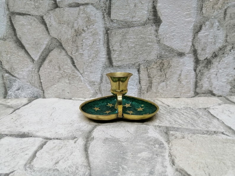May include: A brass candle holder with a green enamel base. The base is shaped like two connected ovals and is decorated with gold stars. The candle holder is on a stone surface.