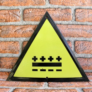 May include: Yellow triangular warning sign with black border and black symbols. The sign has five black plus signs and three black horizontal lines.