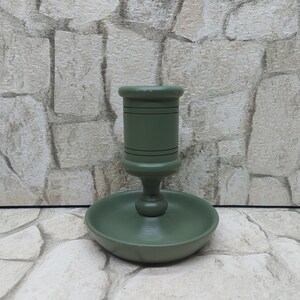 May include: A sage green wooden candlestick holder with a round base and a central cup-shaped holder. The candlestick has a classic design with a slightly distressed finish, suitable for a standard-sized candle. The holder is approximately 15 cm tall.