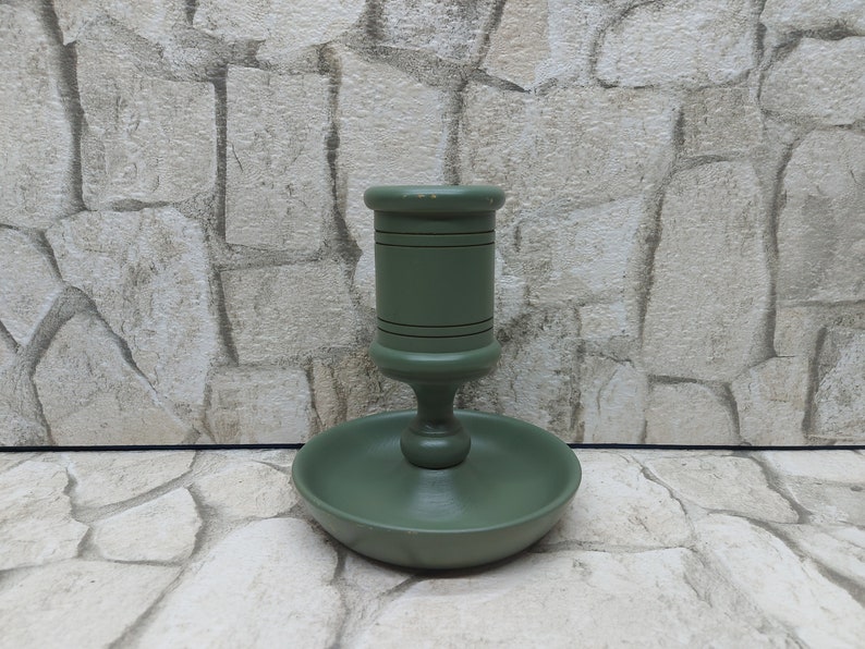 May include: A sage green wooden candlestick holder with a round base and a central cup-shaped holder. The candlestick has a classic design with a slightly distressed finish, suitable for a standard-sized candle. The holder is approximately 15 cm tall.