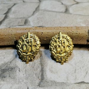 May include: Two gold-colored metal pins with a sword and the letters "DJF" surrounded by a wreath of leaves.