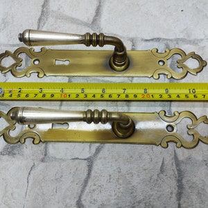 Peut inclure: Deux poignées de porte anciennes en laiton avec plaques arrière ornées. Chaque poignée a un levier argenté et mesure environ 28 cm de long. Les poignées sont présentées sur un fond de briques gris clair.