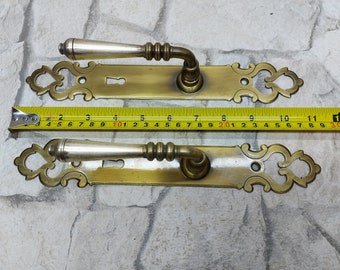 May include: Two antique brass door handles with ornate backplates. Each handle has a silver-colored lever and measures approximately 11 inches in length. The handles are displayed on a light gray brick background.