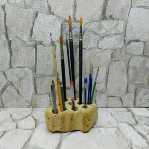 May include: A beige, porous stone brush holder filled with various paint brushes and pens. The brushes have black, wooden, and yellow handles with different bristle shapes. A silver pen and a red and white striped pen are also visible.